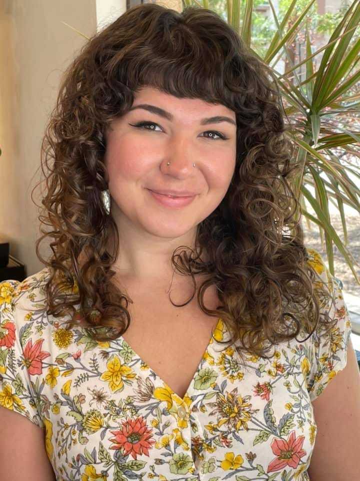 Woman with curly hair and floral blouse smiling in a brightly lit room with plants in the background.