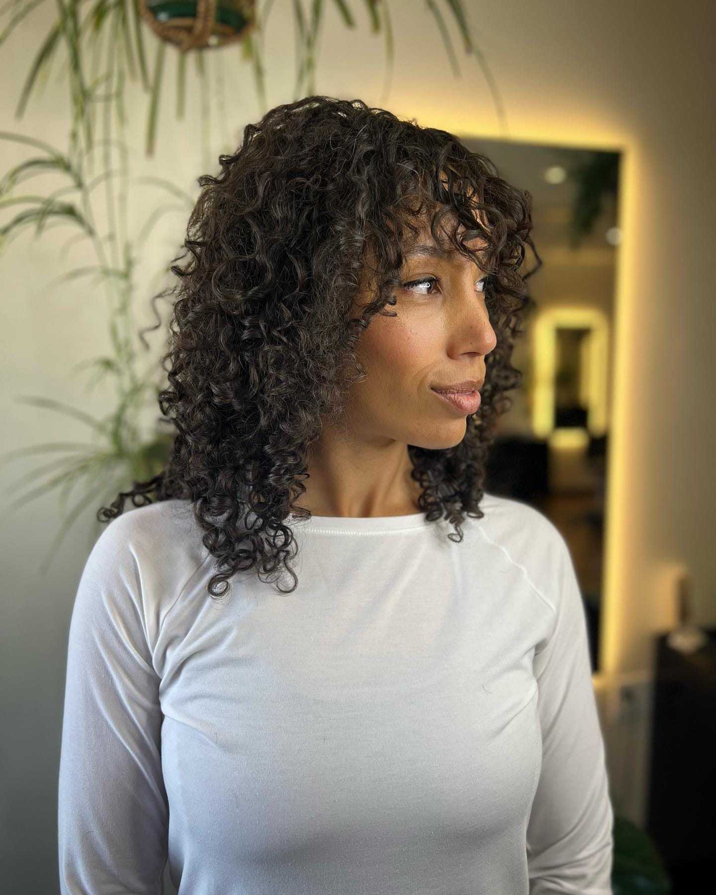 Woman with curly hair in white shirt standing indoors, plants and soft lighting in the background.