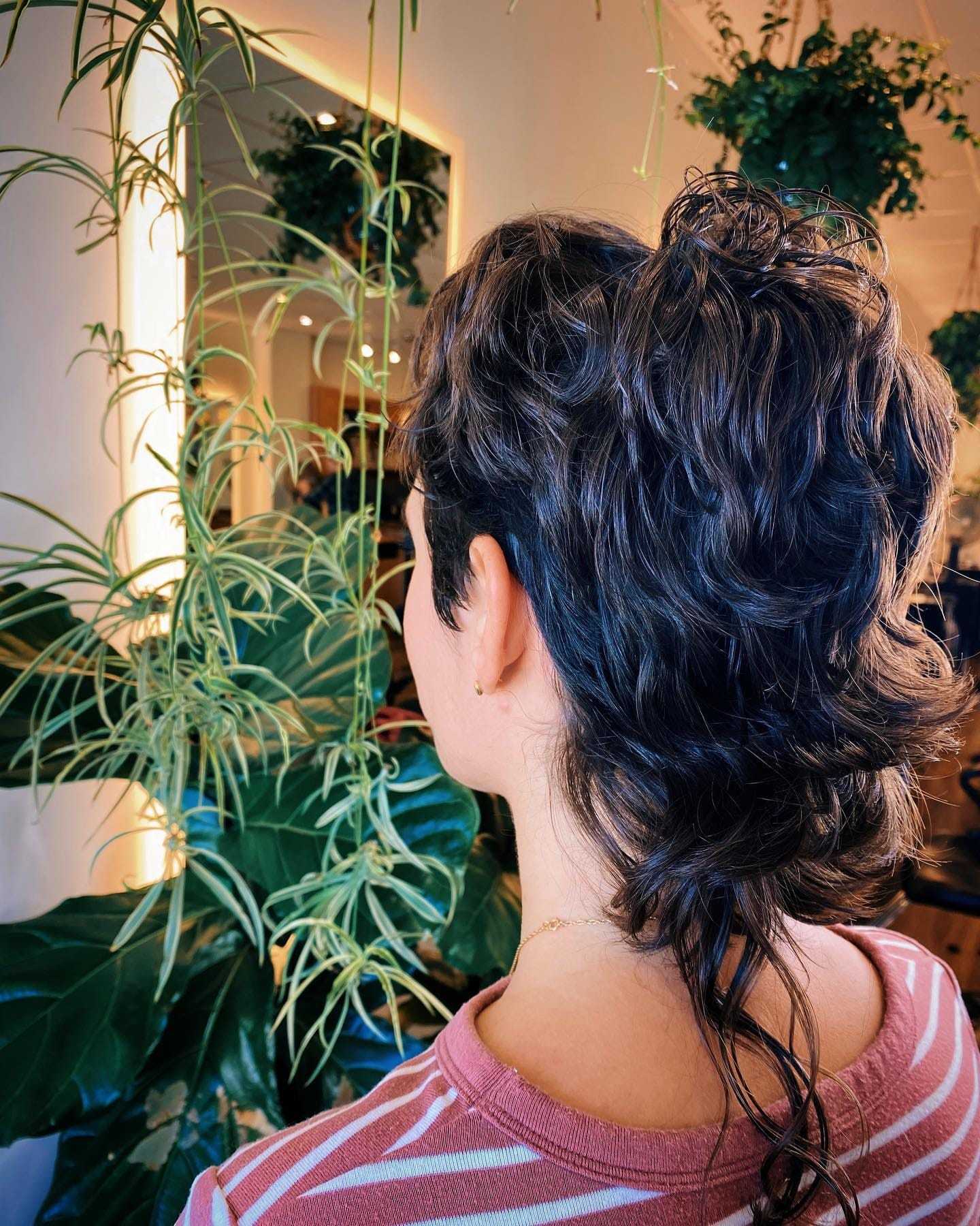 Person with a mullet hairstyle stands near potted plants in a well-lit room.