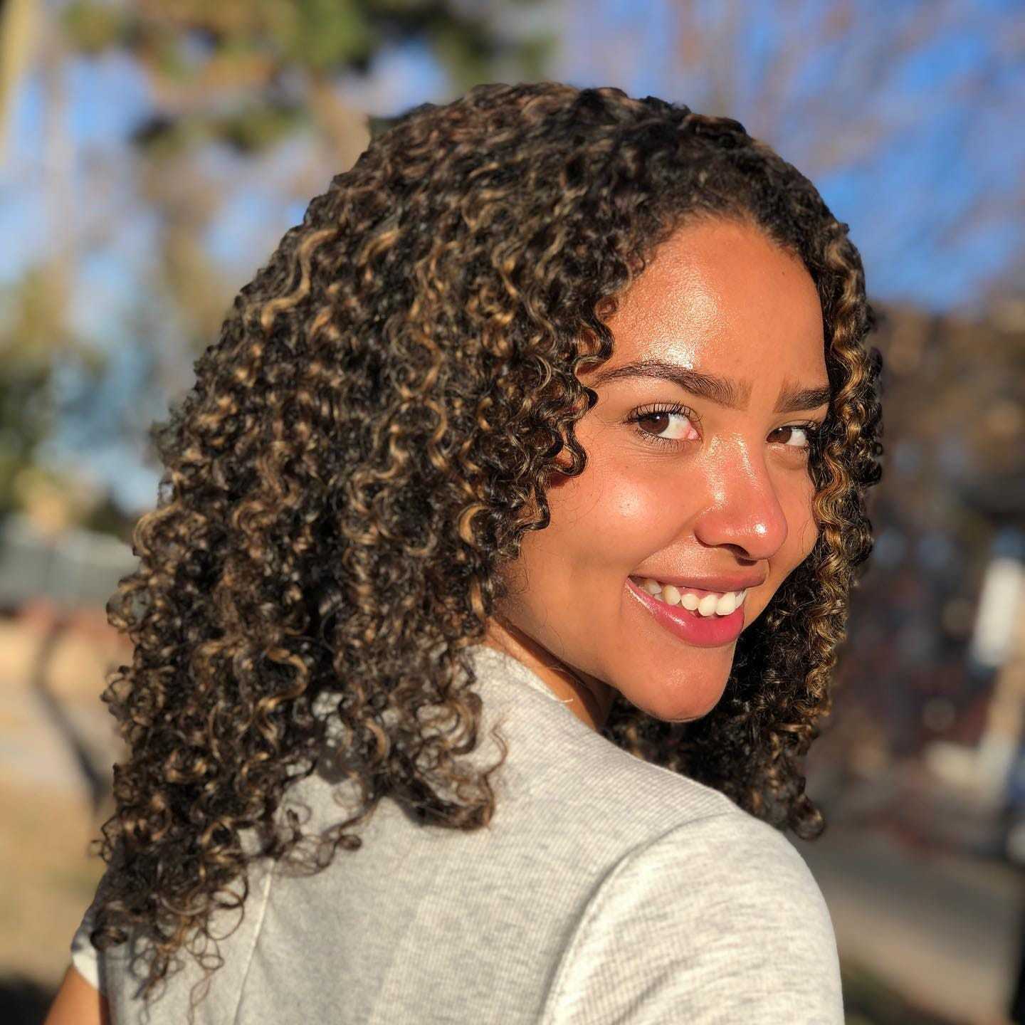 Woman with curly hair smiling in sunlight, wearing a gray shirt, outdoor background blurred.